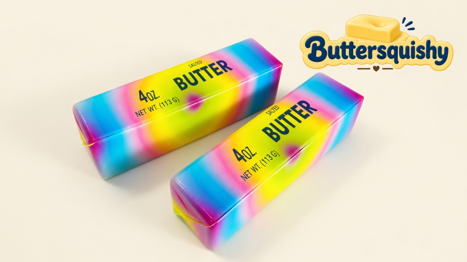 Two Candy Gradient Butter sticks with cyan-to-pink-to-yellow candy melts and navy SALTED BUTTER type on a light-yellow cream studio surface, Buttersquishy logo in the upper right