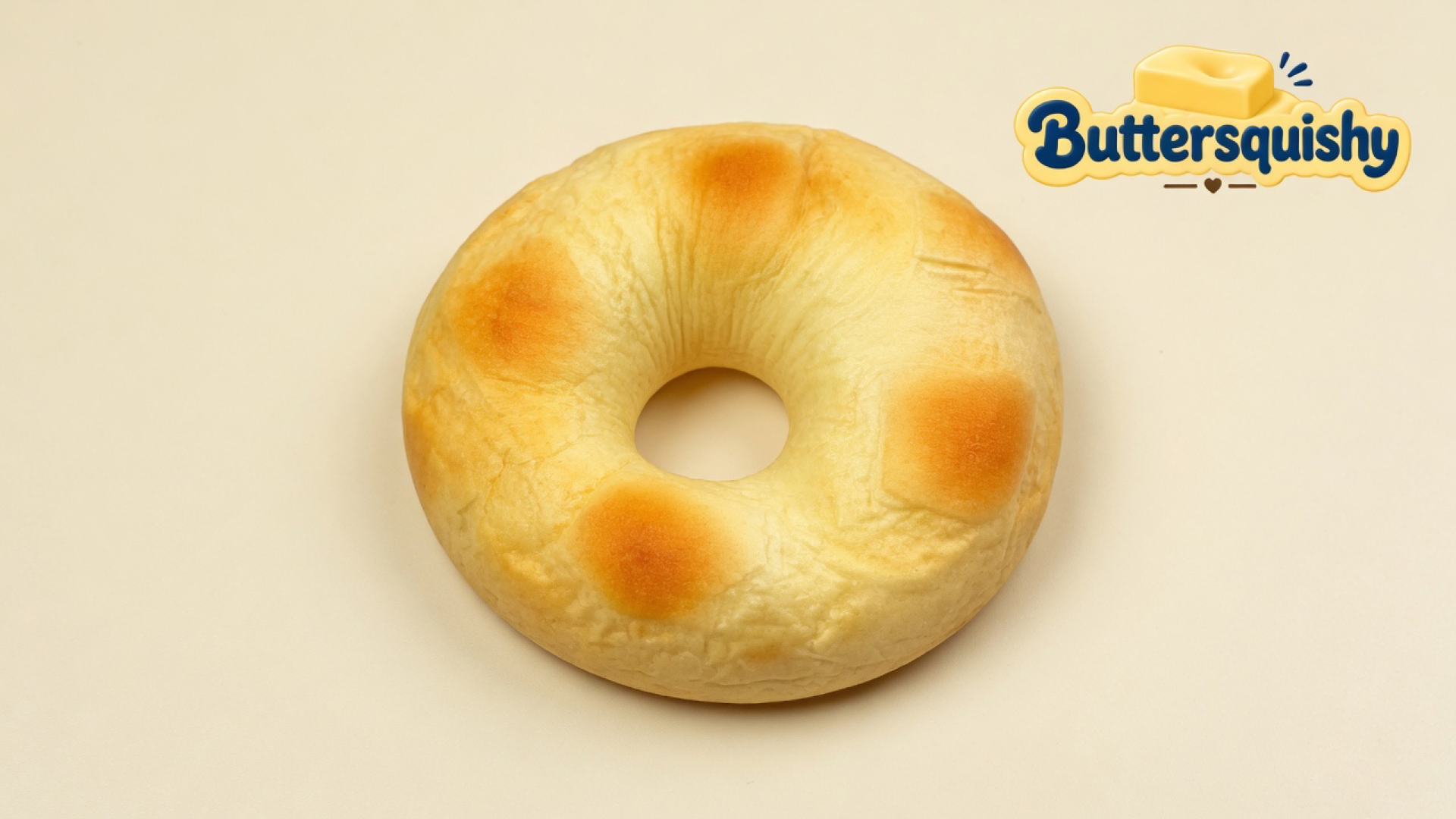 Golden-brown ring-shaped Bread Toast Loaf bun with toasted patches and a center hole on a light-yellow cream studio surface, Buttersquishy logo in the upper right