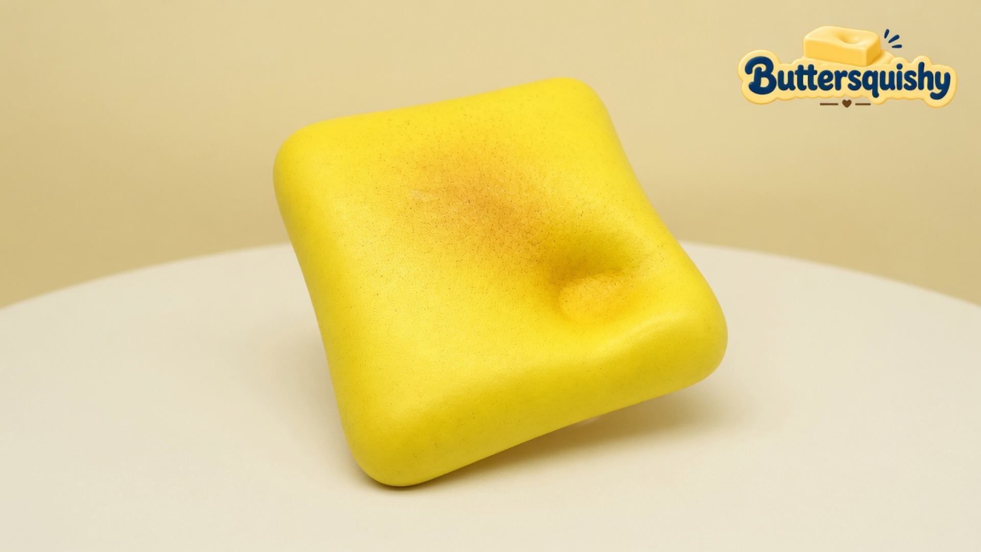 A square butter-yellow Bread Toast Loaf with a toasted-brown center and a thumb dent on a light-yellow cream studio table, Buttersquishy logo in the upper right
