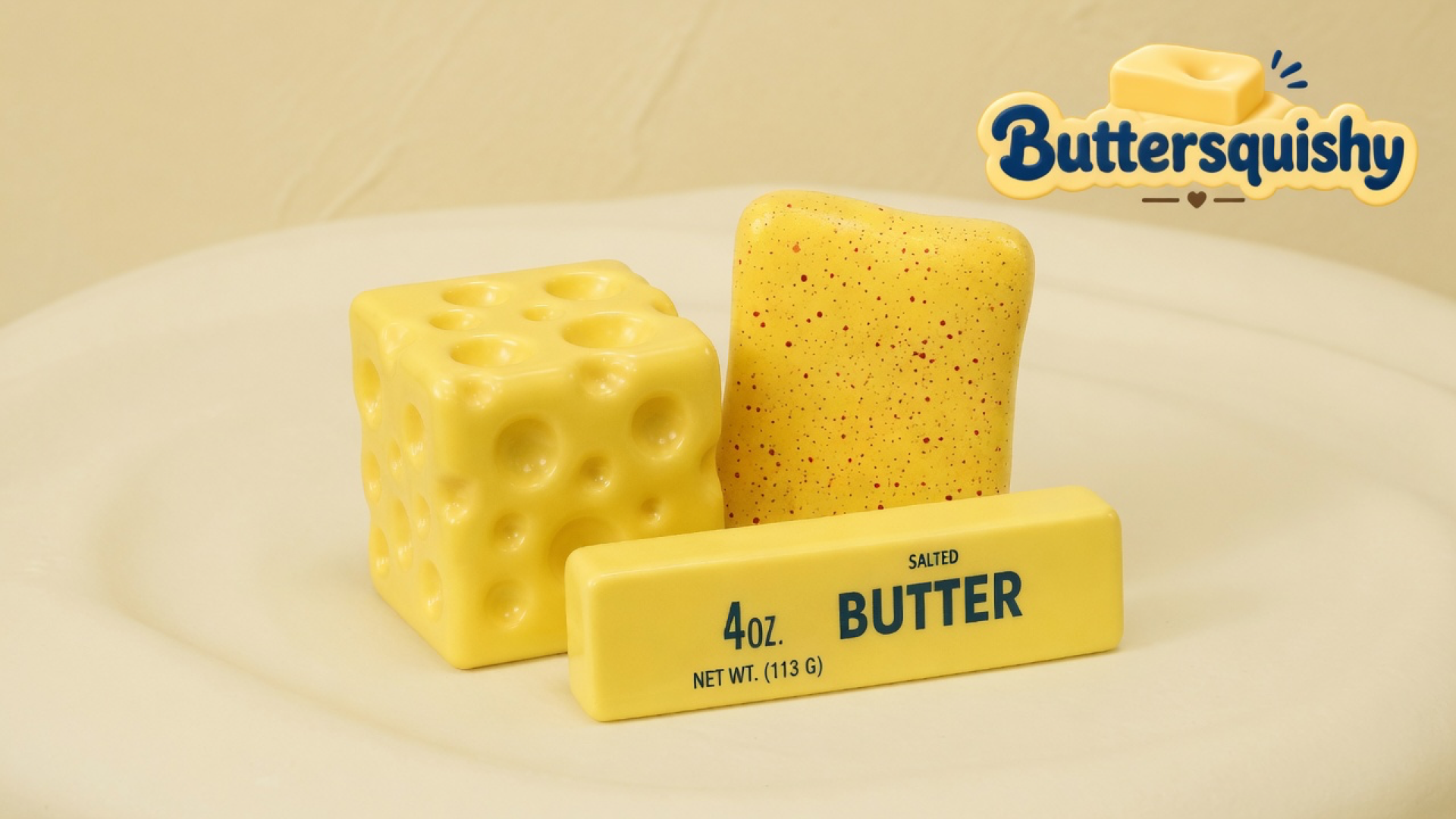 Yellow Swiss-hole cube, red-speckled toast slice, and a 4OZ SALTED BUTTER stick on a cream studio surface, Buttersquishy logo in the upper right