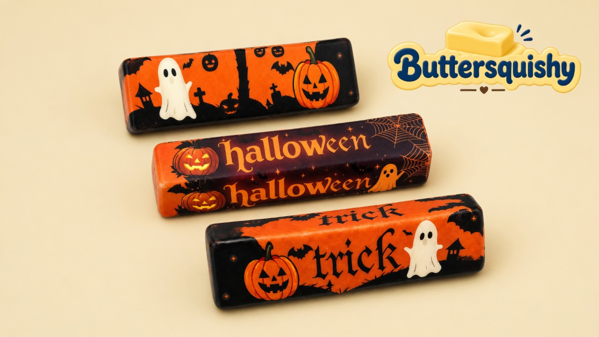 Three Halloween-printed butter sticks — cemetery ghost wrap, stacked halloween lettering, and trick lettering — on a light-yellow cream studio, Buttersquishy logo at upper right