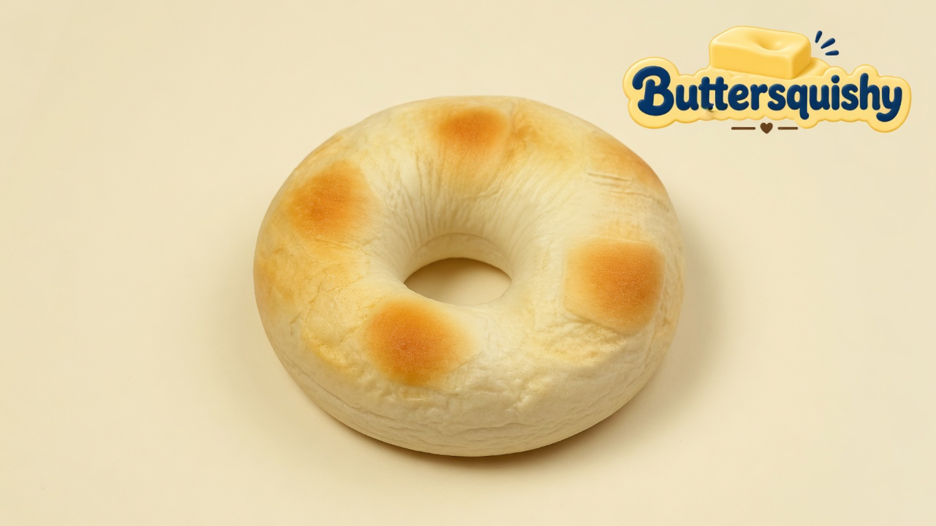 A pale toasted ring loaf with four golden-brown patches and a center hole on a light-yellow cream studio surface, Buttersquishy logo in the upper right