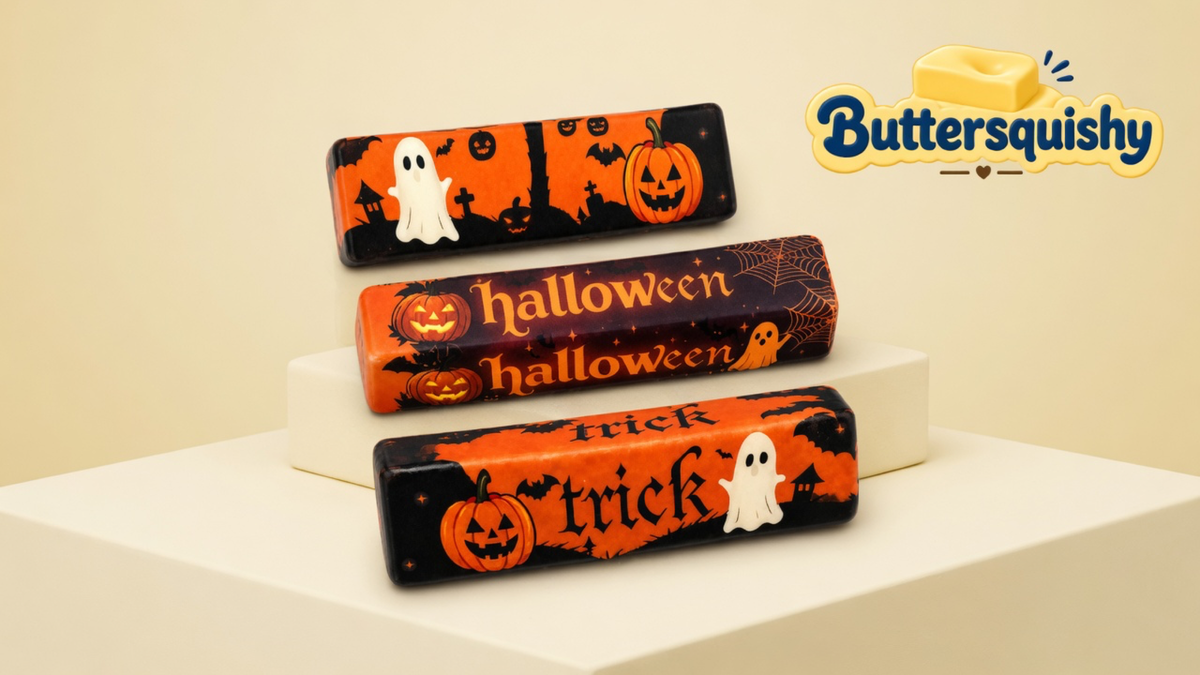 Three Halloween Ghost Butter sticks with a graveyard ghost print, stacked halloween lettering, and stacked trick lettering on a light-yellow cream studio platform, Buttersquishy logo in the upper right