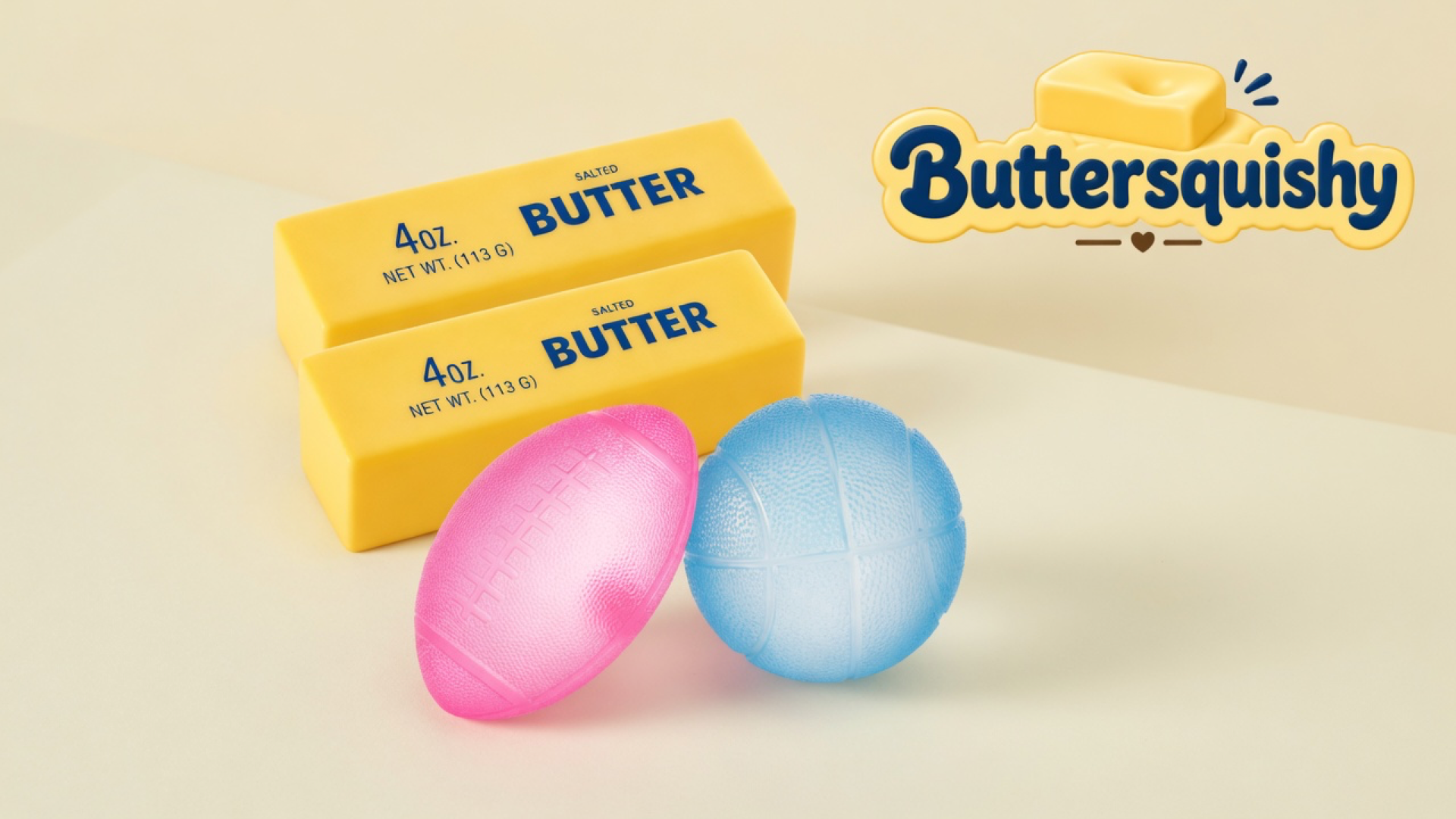 Two stacked yellow salted-butter sticks with a pink football and a pale-blue basketball on a light-yellow cream studio table, Buttersquishy logo in the upper right